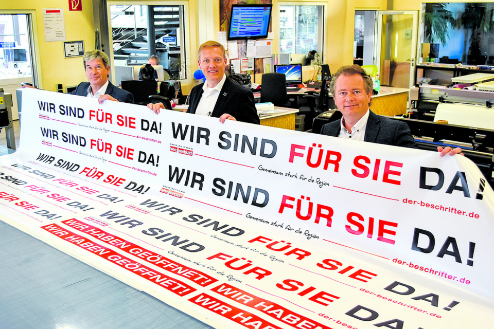 Gemeinsam Flagge zeigen: Holger Grundt (v. li.) und Philipp Brombach von Der Beschrifter sowie Stephan Schrader vom WOCHENBLATT stellen Unternehmen kostenlos Werbebanner zur Verfügung [Foto: ah]